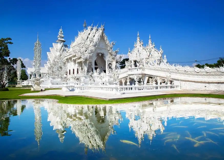 Uno splendido tempio bianco con intricati intagli e guglie si riflette in uno stagno limpido sotto un cielo blu brillante, circondato da una vegetazione lussureggiante: una tappa imperdibile del vostro Tour Tailandia 2026-27 prima di dirigervi verso la vibrante Phuket.