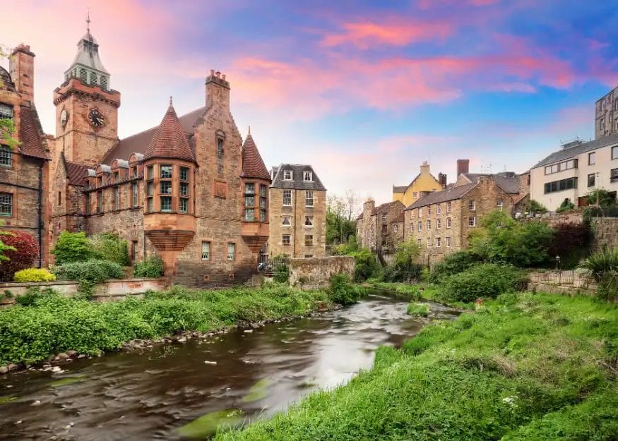 Una vista panoramica di edifici storici in pietra lungo un piccolo fiume in Scozia 2026, con una vegetazione lussureggiante lungo le rive e un cielo colorato al tramonto. L'architettura è caratterizzata da torri dell'orologio e tetti a spioventi, perfetti per il vostro Gran Tour Scozia.
