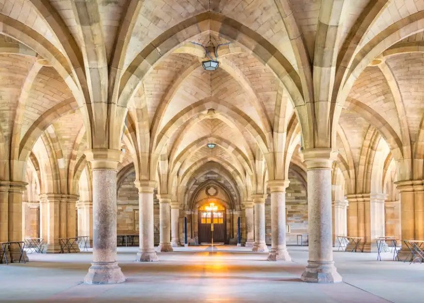 Ampia sala con soffitti ad arco e colonne in pietra, che creano un motivo simmetrico che ricorda una fermata del Gran Tour Scozia. Una luce calda filtra da una porta in fondo, illuminando l'architettura in pietra e le file di sedie allineate ai lati.