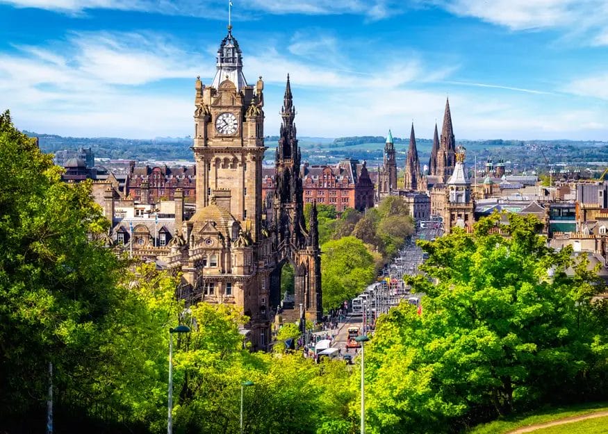 Una vista panoramica di Edimburgo, in Scozia, dal Gran Tour Scozia, con edifici storici in pietra, la torre dell'orologio del Balmoral Hotel, le guglie delle chiese, le strade trafficate e gli alberi verdi sotto un cielo azzurro.