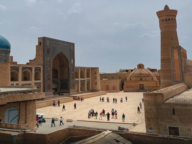 Un gruppo di turisti durante un tour classico della Via della Seta attraversa un'ampia piazza illuminata dal sole in Uzbekistan, circondata da antichi edifici in mattoni: un'imponente struttura a cupola, un ingresso ad arco, una cupola di piastrelle blu e un minareto imponente.
