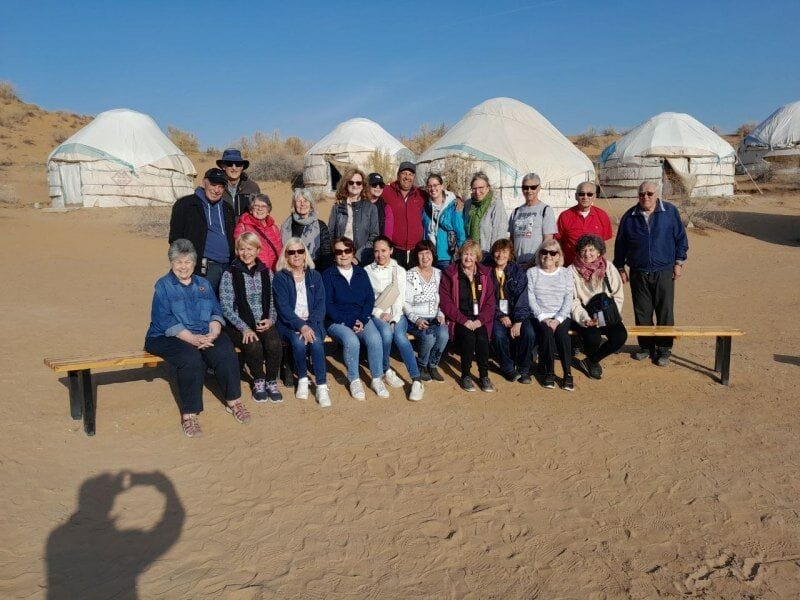 Un gruppo di persone posa per una foto su panchine in un deserto sabbioso vicino al lago Aydarkul, con tende yurta bianche di un Caravan tour Uzbekistan e arbusti secchi sullo sfondo, sotto un cielo azzurro e limpido. L'ombra del fotografo è visibile di fronte.