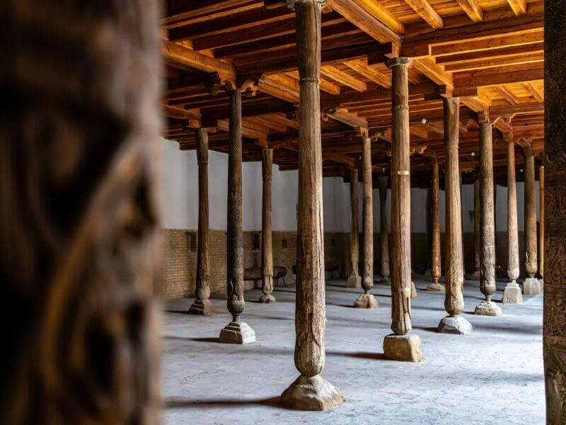 Una sala spaziosa con numerose colonne in legno intagliato che sostengono un soffitto in legno, creando disegni di luci e ombre sul pavimento in pietra. La scena tranquilla e storica evoca il patrimonio della Via della Seta dell'Uzbekistan, con le sue pareti beige che ricordano un classico tour nel tempo.