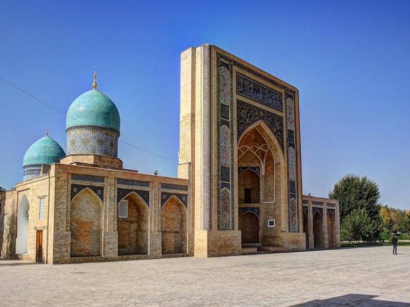 Una moschea storica con due grandi cupole turchesi e un ingresso alto e decorato, con intricate piastrelle e pareti in mattoni, sullo sfondo di un cielo azzurro e limpido, perfetto da ammirare durante un tour in carovana dell'Uzbekistan o un campeggio yurta vicino al lago Aydarkul.