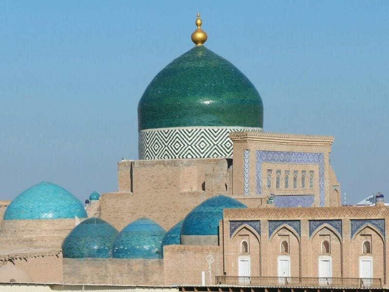 Una grande cupola turchese con intricati motivi geometrici e diverse cupole blu più piccole coronano un edificio storico in mattoni con finestre ad arco e piastrelle a mosaico: una vista iconica in qualsiasi tour classico della Via della Seta dell'Uzbekistan, sullo sfondo di un cielo azzurro e limpido.