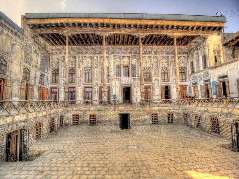 Grande edificio storico con balcone in legno ornato, motivi dettagliati, finestre ad arco e un ampio cortile in pietra: perfetto per un'avventura in carovana in Uzbekistan prima di rilassarsi accanto al lago Aydarkul o provare il campeggio yurta sotto il cielo parzialmente nuvoloso.