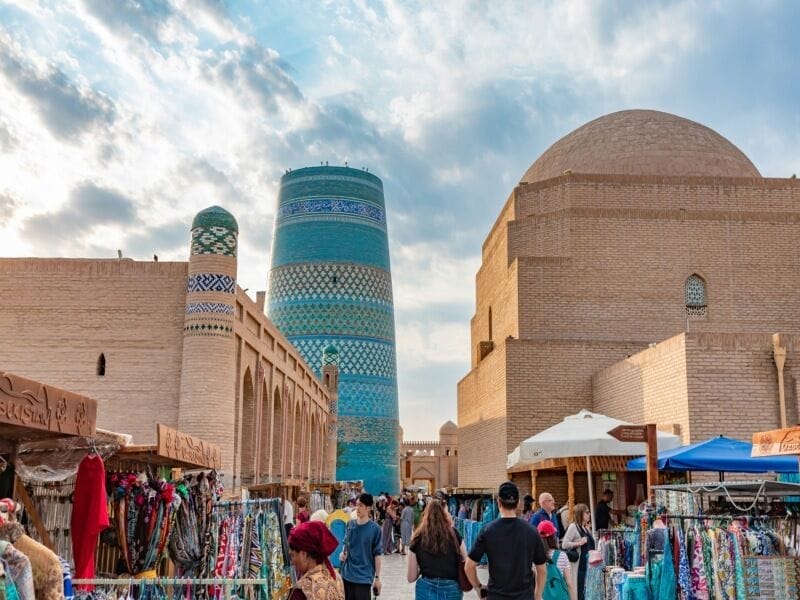 Un vivace mercato con persone che curiosano tra le bancarelle colorate si staglia contro gli edifici storici in mattoni e il minareto turchese Kalta Minor a Khiva, in Uzbekistan, una tappa iconica lungo la leggendaria Via della Seta.