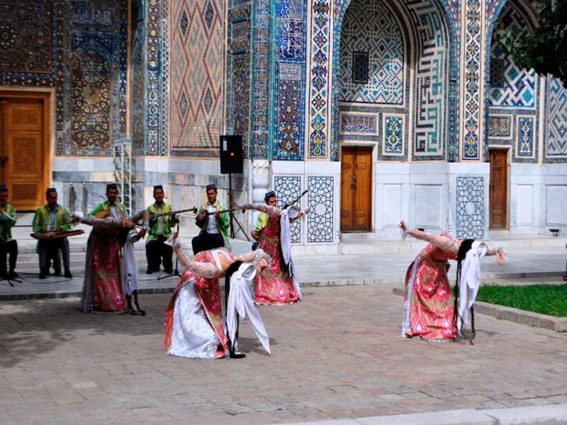 Quattro donne in costumi tradizionali colorati eseguono una danza all'aperto, mentre musicisti in verde suonano davanti a un edificio ornato di piastrelle, catturando lo spirito vibrante di un tour in carovana in Uzbekistan vicino al campeggio yurta sul lago Aydarkul.