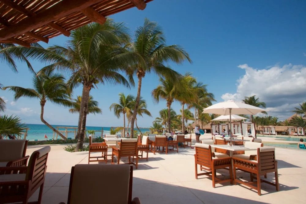 Area salotto all'aperto di un resort termale sulla spiaggia con tavoli in legno e sedie imbottite, palme, ombrelloni e cielo azzurro con vista sulla baia di Akumal e sulla spiaggia sabbiosa sullo sfondo.