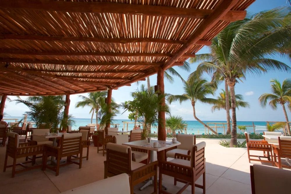 Ristorante all'aperto con tavoli e sedie in legno sotto una pergola, con vista sulla spiaggia tropicale della baia di Akumal, con palme e oceano blu: una cornice perfetta per un pasto rilassante in questo resort sulla spiaggia sotto un cielo limpido.