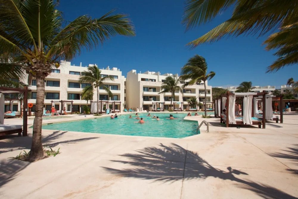 Le persone nuotano e si rilassano in una grande piscina all'aperto circondata da palme e cabanas presso l'Akumal Bay Beach Resort, con un moderno edificio alberghiero a più piani sullo sfondo, sotto un cielo azzurro e limpido.