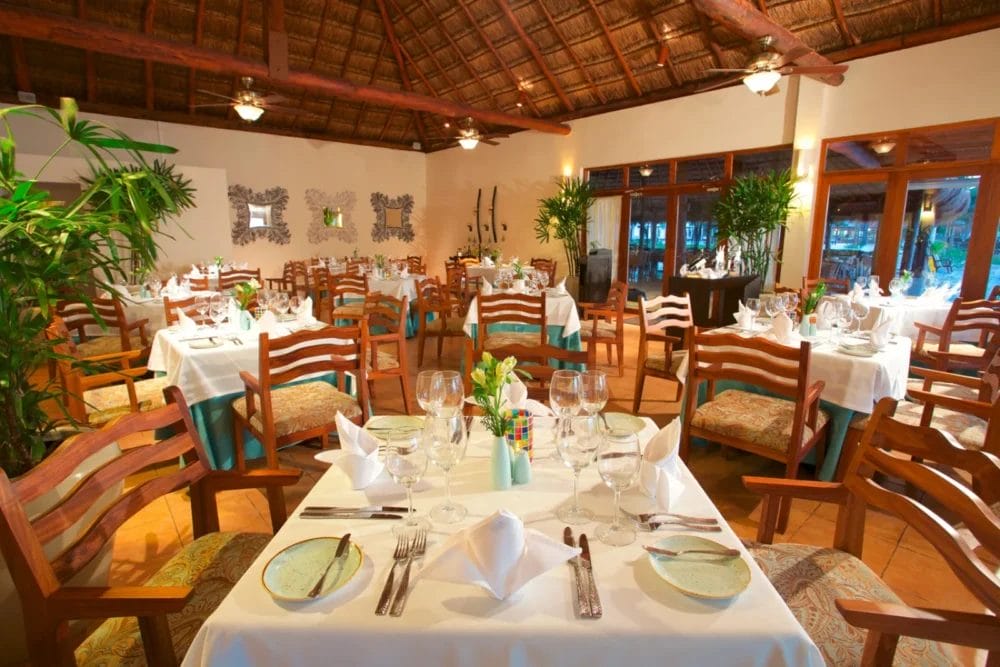 Lo spazioso ristorante di questo resort balneare dispone di sedie e tavoli in legno con tovaglie, bicchieri e stoviglie bianche. Grandi finestre, piante verdi e un tetto di paglia creano un'atmosfera calda e invitante vicino alla baia di Akumal.