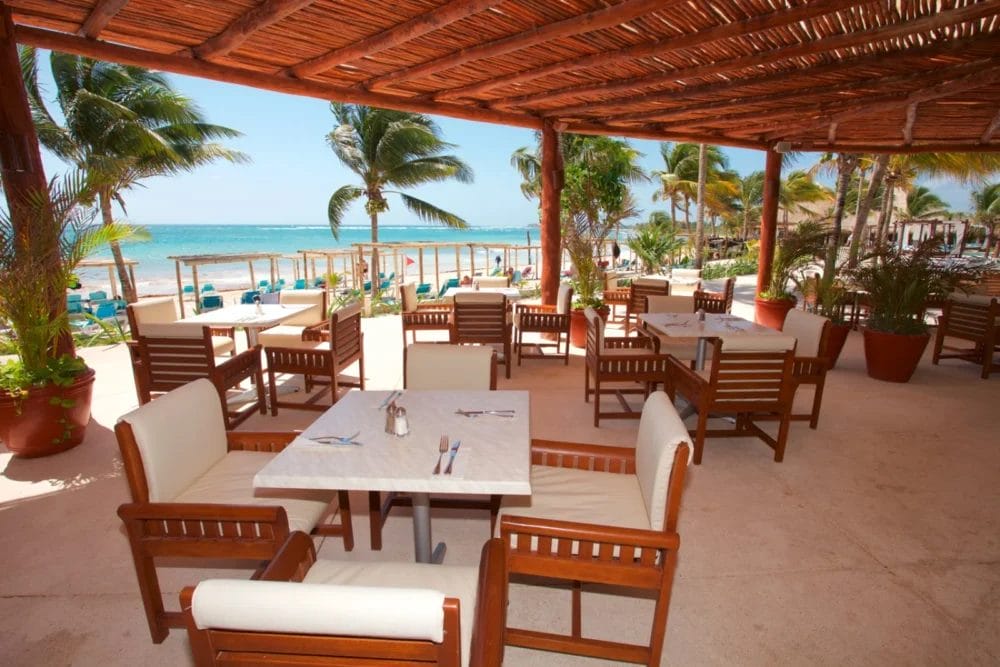 Il patio del ristorante all'aperto, con tavoli in legno e sedie imbottite, si affaccia sulla spiaggia sabbiosa e sulle palme della baia di Akumal, all'ombra di un tetto in legno, creando una rilassante atmosfera tropicale in questo invitante resort sulla spiaggia.