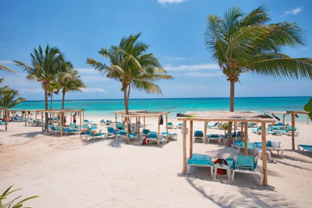 Spiaggia sabbiosa con palme, sedie a sdraio blu e cabine in legno che si affacciano sull'acqua turchese dell'oceano, sotto un cielo limpido e azzurro, in questo rilassante resort sulla spiaggia di Akumal Bay. Le persone si godono l'ambiente soleggiato e tropicale.