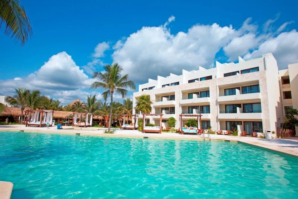 Un resort tropicale sulla spiaggia della baia di Akumal con una grande piscina, sedie a sdraio, palme e un moderno edificio bianco sotto un cielo blu con nuvole soffici.