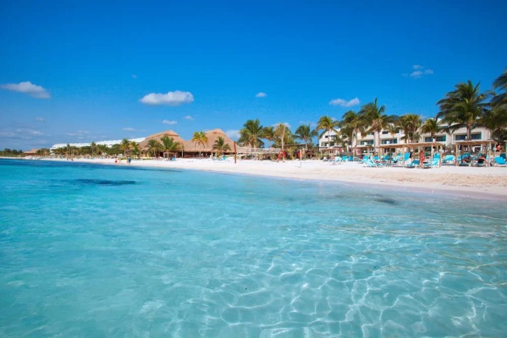L'acqua limpida e turchese conduce a una spiaggia sabbiosa fiancheggiata da palme, sedie a sdraio ed edifici del Beach Resort sotto un cielo blu brillante con qualche nuvola sparsa.