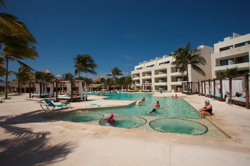 Un'area della piscina del resort con persone che nuotano e si rilassano al sole. Moderni edifici bianchi, palme e cabanas circondano la grande piscina di questo resort termale della baia di Akumal sotto un cielo azzurro e limpido.