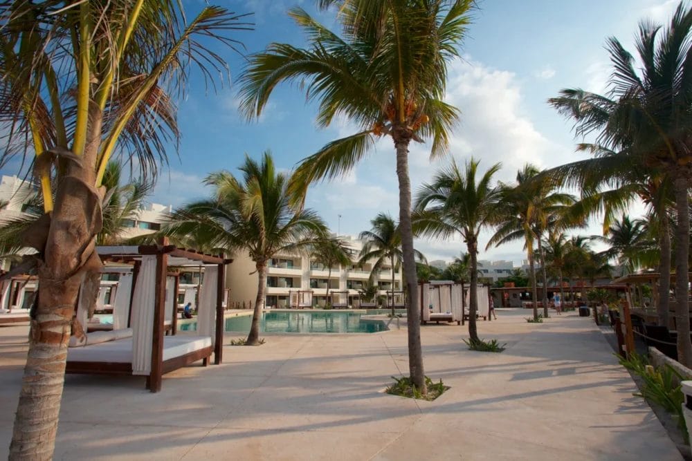 Una splendida area piscina di un resort sulla spiaggia con palme, cabanas bianche con letti e moderni edifici a più piani sullo sfondo, sotto un cielo azzurro parzialmente nuvoloso, che cattura l'atmosfera rilassante della baia di Akumal.