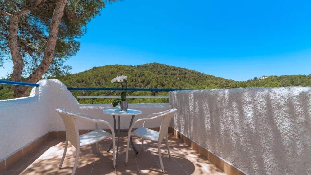 Un piccolo patio del Globales Montemar è caratterizzato da due sedie di vimini bianche e da un tavolo rotondo con un'orchidea in vaso, che si affaccia su colline verdi sotto un cielo azzurro e limpido, il tutto incorniciato da pareti di stucco bianco e alberi.