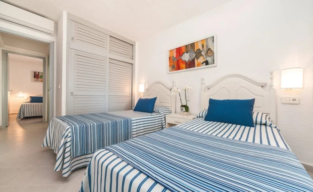 Una luminosa camera da letto dell'HOTEL GLOBALES MONTEMAR presenta due letti singoli con lenzuola a righe bianche e blu, cuscini blu, un comodino con un'orchidea bianca, lampade a muro, armadi a muro e un dipinto colorato sopra i letti.