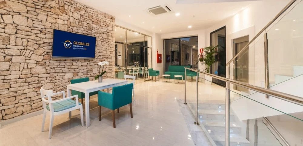 Lobby moderna e ben illuminata nello stile dell'HOTEL GLOBALES MONTEMAR, con parete in pietra, grande display digitale, sedie verde acqua, tavoli bianchi, pareti in vetro e scala con ringhiera in acciaio inox. Sono visibili piante da interno e un estintore.