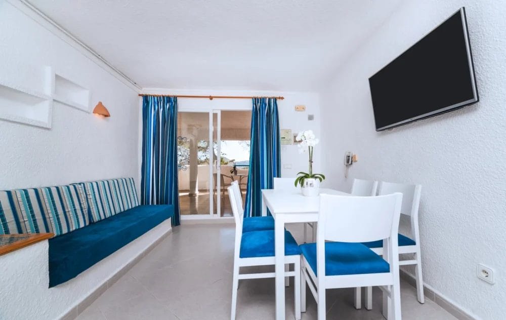 La luminosa e moderna zona pranzo dell'HOTEL GLOBALES MONTEMAR presenta un tavolo bianco e quattro sedie con cuscini blu, una panca a righe blu, una TV a parete e porte scorrevoli in vetro che conducono a un balcone con posti a sedere all'aperto.
