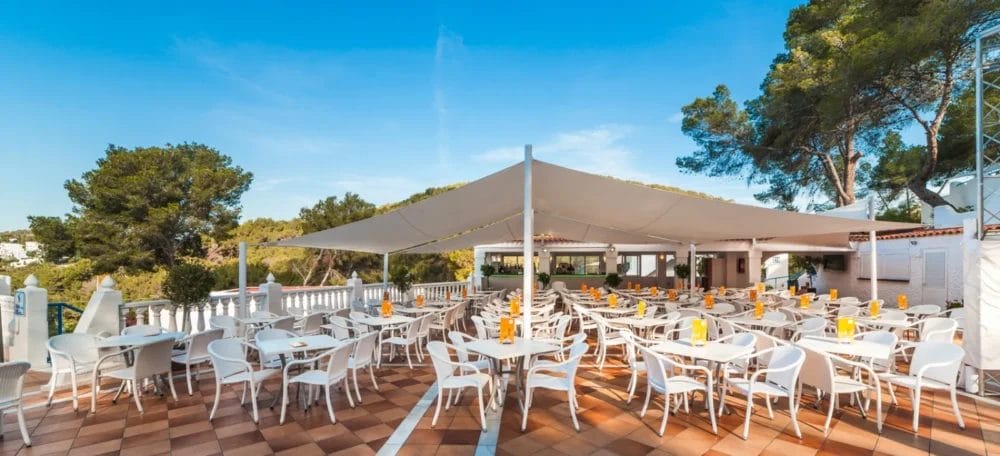 La terrazza del ristorante all'aperto dell'HOTEL GLOBALES MONTEMAR, con tavoli e sedie bianchi disposti ordinatamente sotto un'ampia tettoia, circondata da alberi e verde sotto un cielo azzurro e limpido.