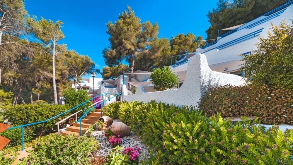 Una scena soleggiata all'aperto presso l'HOTEL GLOBALES MONTEMAR, con edifici dalle pareti bianche con ringhiere blu, piante verdi, fiori colorati e alti pini sotto un cielo azzurro. I gradini salgono attraverso il vivace giardino.