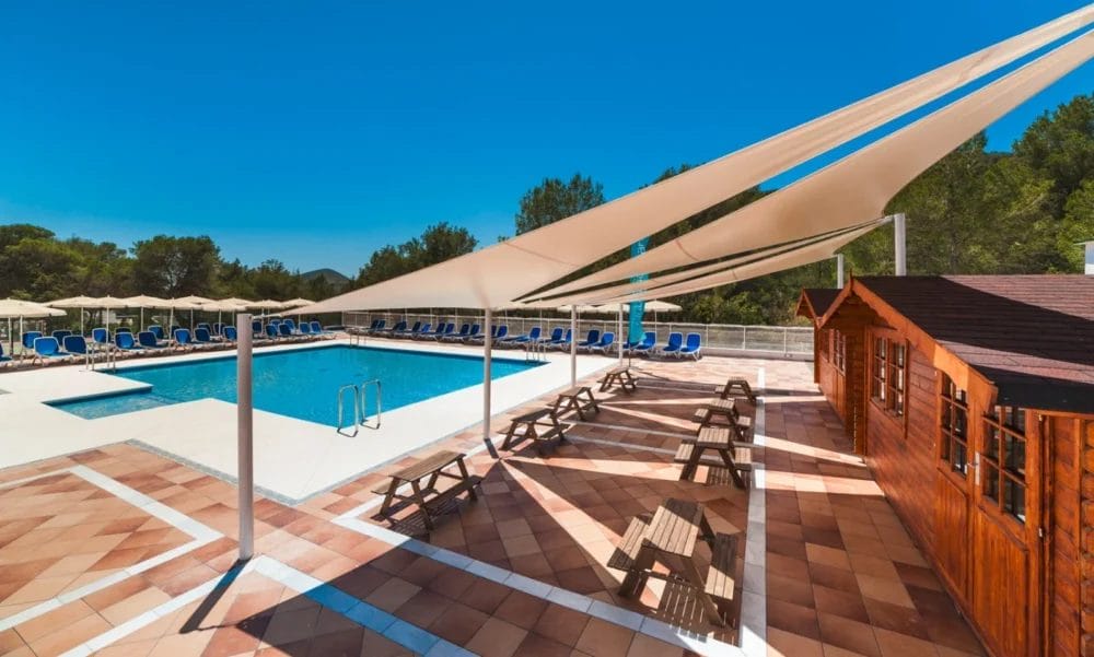 Piscina all'aperto circondata da sedie a sdraio e ombrelloni all'HOTEL GLOBALES MONTEMAR, con vele ombreggianti triangolari sopra tavoli da picnic su un patio piastrellato; un edificio in legno è sulla destra, mentre alberi e cielo blu completano la scena.