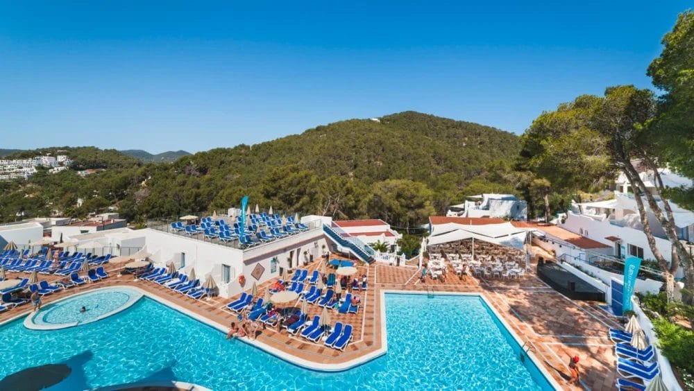Un'ampia area della piscina all'aperto del resort collinare HOTEL GLOBALES MONTEMAR, con sedie a sdraio blu che circondano due piscine. Sullo sfondo edifici bianchi, alberi verdi e colline boscose, sotto un cielo azzurro e limpido.