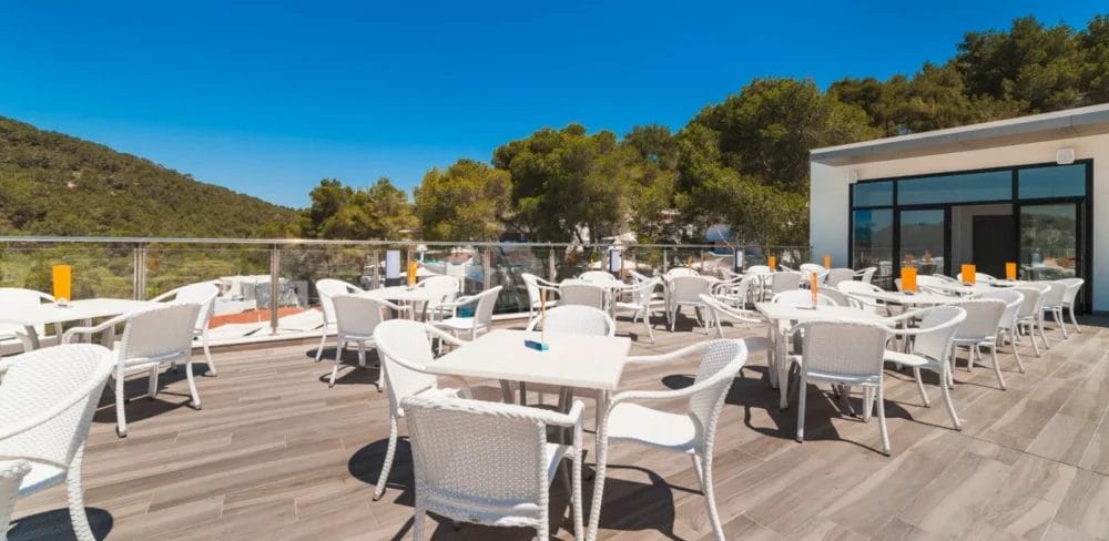 Patio del ristorante all'aperto dell'HOTEL GLOBALES MONTEMAR con tavoli e sedie bianche su una terrazza di legno, circondata da una ringhiera di vetro. Il verde degli alberi e delle colline fa da sfondo a un cielo azzurro e limpido.