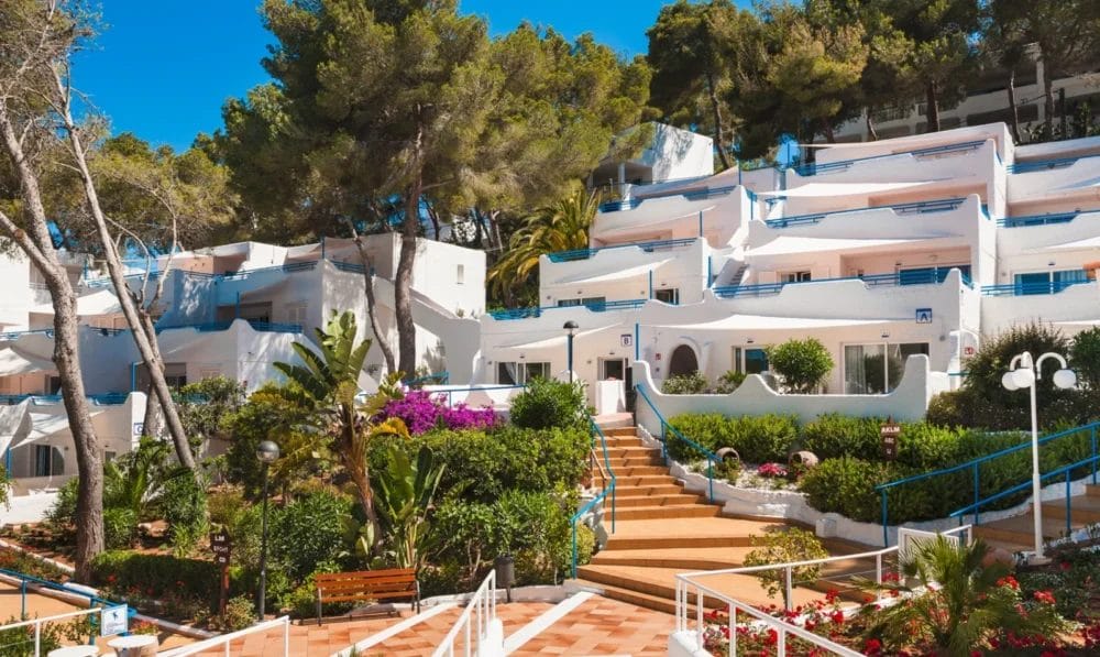 Gli edifici bianchi in stile mediterraneo con ringhiere blu dell'HOTEL GLOBALES MONTEMAR sono circondati da una vegetazione lussureggiante, palme e fiori colorati. Scale e sentieri in terracotta si snodano attraverso i giardini sotto un cielo azzurro.