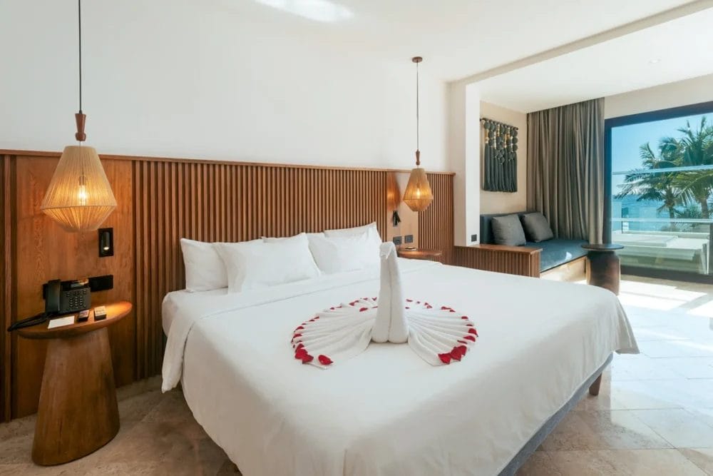 Una moderna camera d'albergo Beach Resort con un grande letto ornato da cigni di spugna e petali di rosa, accenti in legno, luci a sospensione, un'area salotto accanto a una grande finestra e una vista mozzafiato sulle palme della baia di Akumal e sull'oceano esterno.