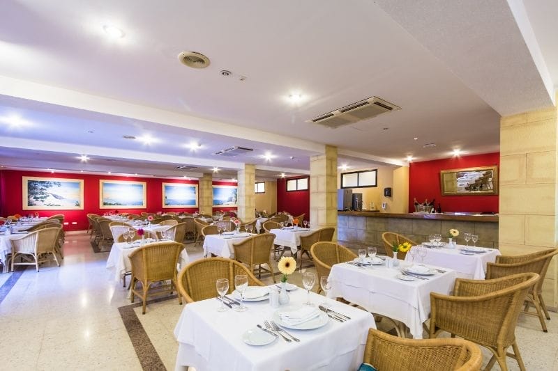Ampio ristorante dell'HOTEL BLUEBAY, con sedie di vimini e tavoli apparecchiati con tovaglie bianche, piatti, bicchieri e fiori. Le pareti rosse presentano foto incorniciate e nell'angolo in fondo a destra c'è un bancone, perfetto per un'esperienza BEACH CLUB in un hotel 4 stelle.