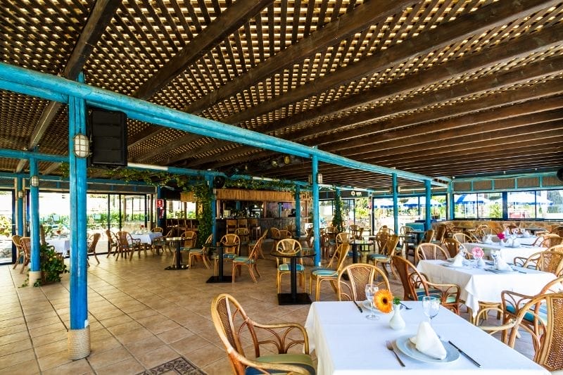 Ristorante all'aperto dell'HOTEL BLUEBAY BEACH CLUB con travi in legno e accenti blu, con sedie in vimini e tovaglie bianche. I tavoli sono apparecchiati con tovaglioli, bicchieri e fiori, mentre le grandi finestre offrono una vista sul verde all'esterno di questo hotel 4 stelle.