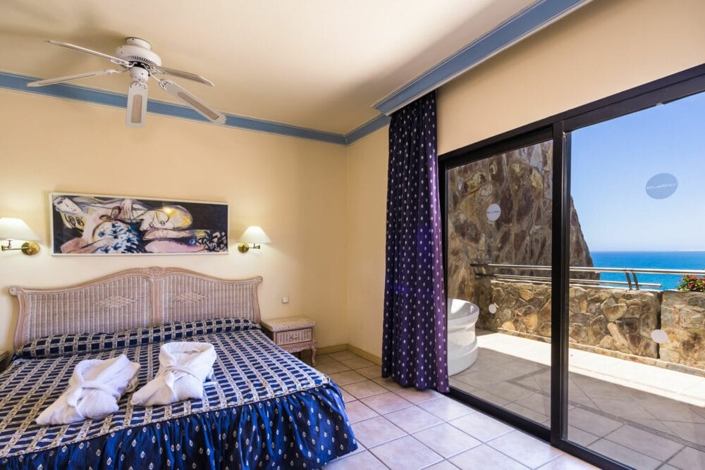 La luminosa camera dell'HOTEL BLUEBAY BEACH CLUB è dotata di un grande letto con lenzuola a fantasia blu, cigni in asciugamano, ventilatore a soffitto e porte scorrevoli in vetro che si aprono su un balcone con una splendida vista sull'oceano e sulle scogliere sotto un cielo limpido.
