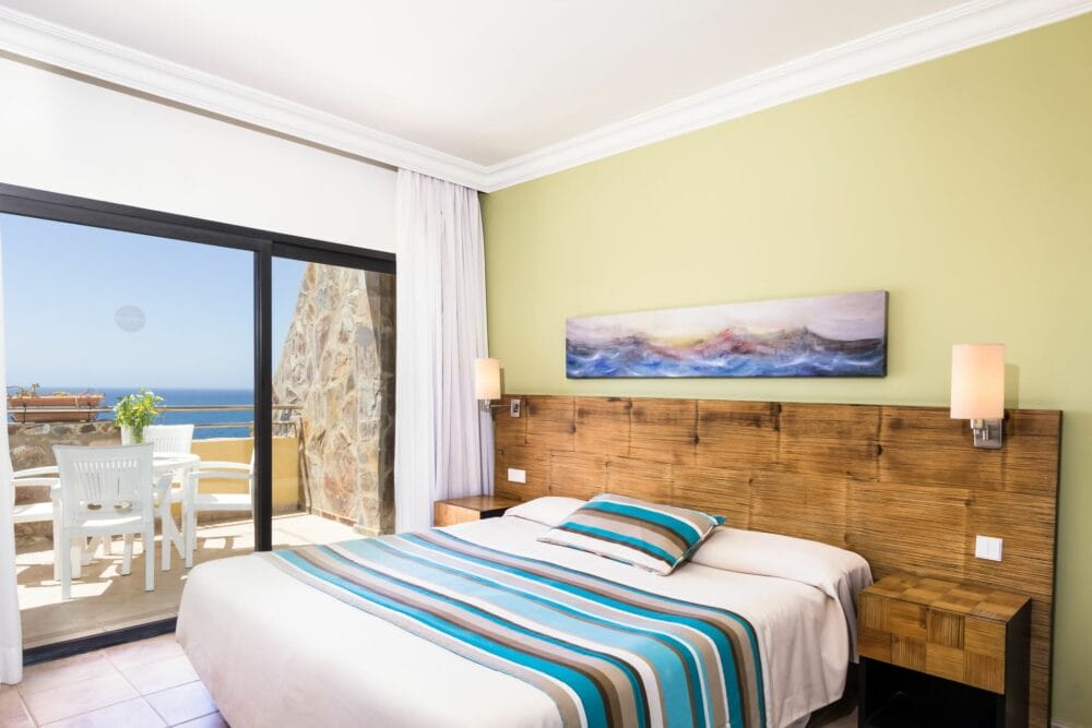 La luminosa camera a 4 stelle dell'HOTEL BLUEBAY è dotata di un grande letto con lenzuola a righe, testiera in legno, comodini, lampade a muro e porte scorrevoli in vetro su un balcone che si affaccia sull'oceano: godetevi lo stile del resort come un vero e proprio BEACH CLUB.