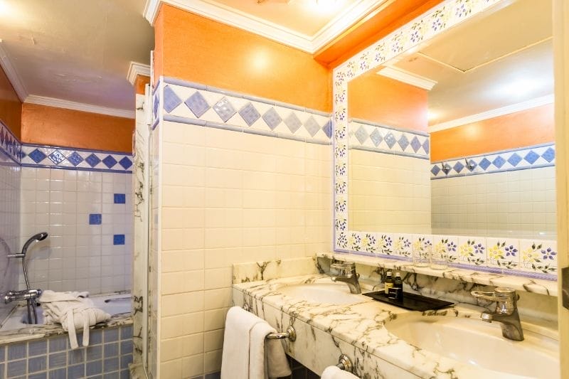 Il bagno dell'hotel 4 stelle HOTEL BLUEBAY BEACH CLUB presenta pareti arancioni e bianche, un piano di lavoro in marmo con doppio lavabo, un grande specchio delimitato da piastrelle floreali e una vasca da bagno con piastrelle blu e bianche. Gli asciugamani sono collocati vicino ai lavandini e alla vasca.