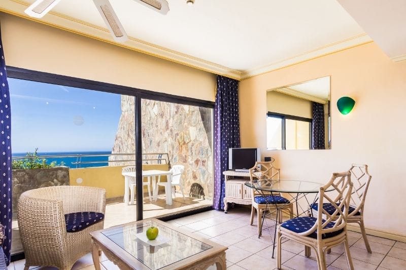 Soggiorno luminoso e moderno dell'hotel 4 stelle HOTEL BLUEBAY con mobili in vimini e vetro, accenti blu, uno specchio a parete e grandi porte scorrevoli che si aprono su un balcone con vista sull'oceano sotto un cielo limpido, perfetto per il relax del BEACH CLUB.