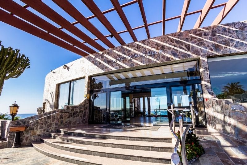 Un moderno edificio in pietra con grandi porte e finestre in vetro, un pergolato in alto che proietta ombre e i gradini che conducono all'ingresso dell'HOTEL BLUEBAY, un hotel a 4 stelle; sullo sfondo, cactus e oceano.