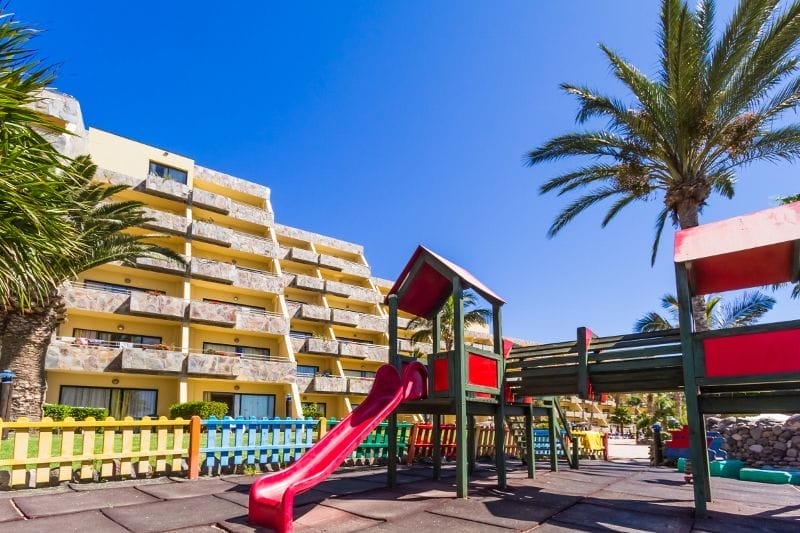 Un parco giochi colorato con scivoli e strutture per l'arrampicata è circondato da palme, accanto all'HOTEL BLUEBAY, un hotel 4 stelle giallo a più piani, sotto un cielo azzurro e limpido.