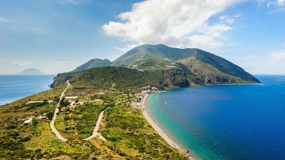 Veduta aerea di un'isola verde e lussureggiante nelle isole eolie, con una strada tortuosa, piccole case, campi terrazzati e una spiaggia di ciottoli curvilinea con acqua azzurra e limpida, sostenuta da montagne sotto un cielo parzialmente nuvoloso. Perfetto per un indimenticabile tour eolie.