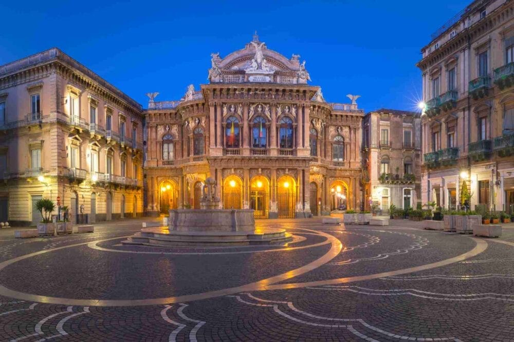 Un teatro storico dall'architettura ornata e dalle luci scintillanti si trova al centro di una piazza acciottolata della Sicilia orientale, circondato da edifici eleganti, sotto un cielo azzurro e limpido, un momento perfetto del vostro tour della Sicilia.