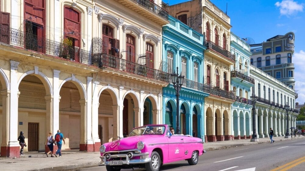 In una strada soleggiata, una decappottabile d'epoca rosa acceso passa accanto a edifici colorati in stile coloniale con archi e balconi vicino alla Casa Particular Classique, mentre diverse persone camminano sul marciapiede.