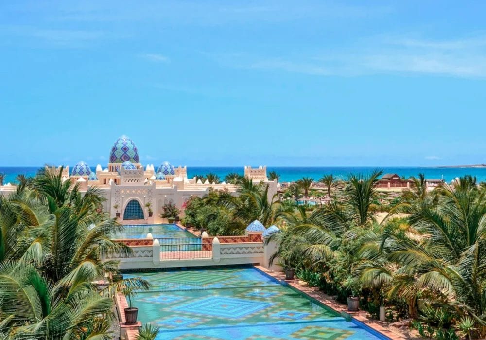 Un grande resort tropicale, il SEARESORT RIU KARAMBOA on BOAVISTA, caratterizzato da tetti blu a cupola, palme, piscine decorate e giardini lussureggianti sullo sfondo del cielo blu e dell'oceano.