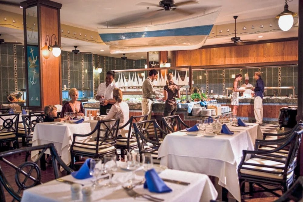 Il ristorante del RIU KARAMBOA a Boavista presenta decorazioni nautiche, tra cui una barca appesa al soffitto. I commensali possono usufruire del servizio a buffet su tavoli con tovaglie bianche e tovaglioli blu, in un'atmosfera da vero SEARESORT.