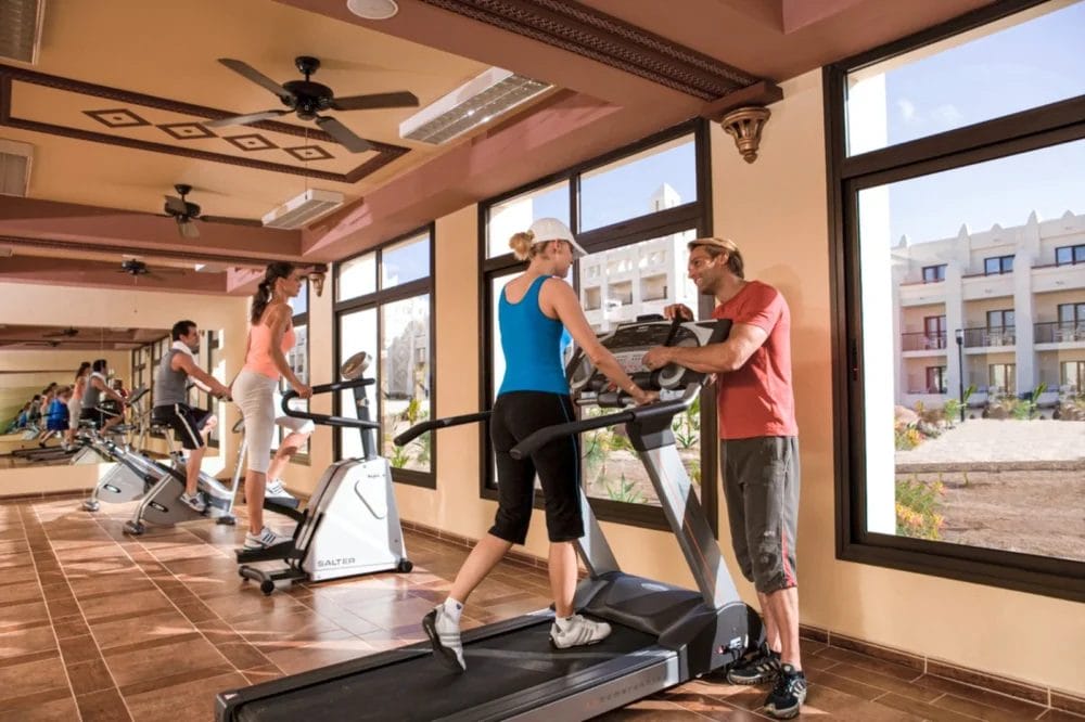 Persone si allenano su tapis roulant in una palestra luminosa, spaziosa e con grandi finestre. Un istruttore parla con una donna su un tapis roulant mentre altre due corrono sullo sfondo. All'esterno si vedono il SEARESORT RIU KARAMBOA e il verde di Boavista.