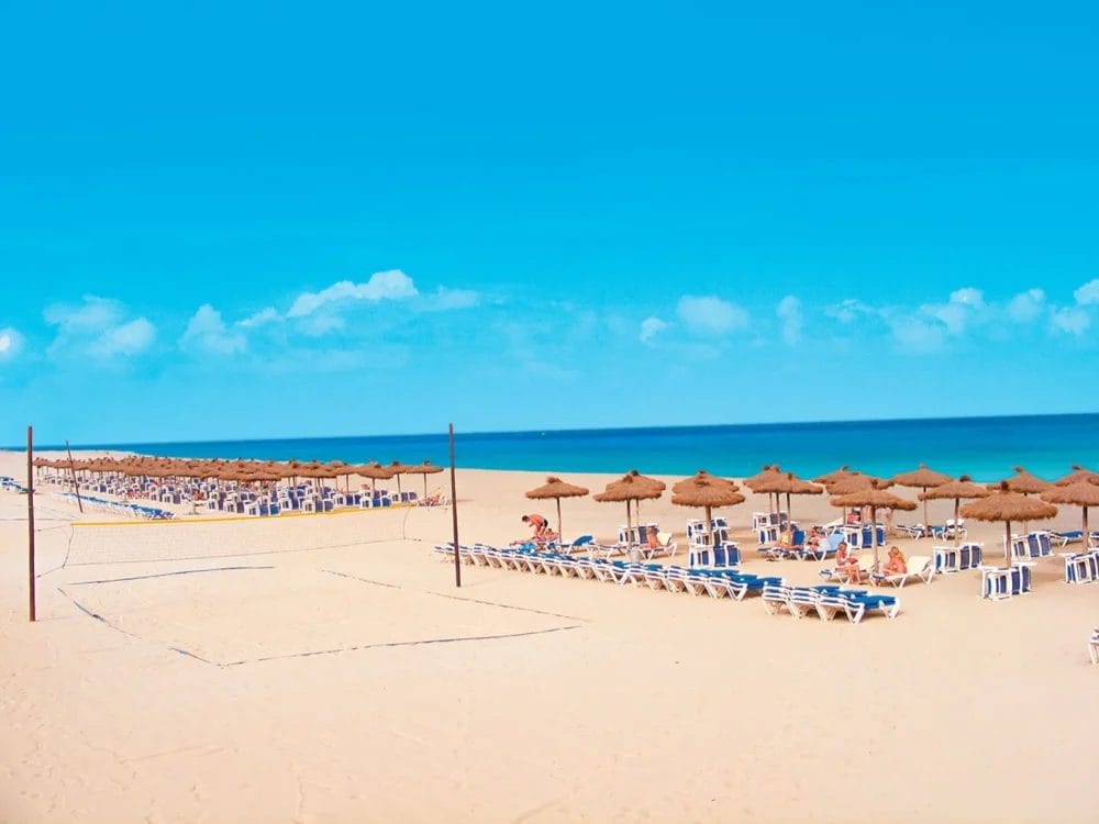 File di sedie a sdraio vuote e ombrelloni di paglia costeggiano la spiaggia sabbiosa del SEARESORT RIU KARAMBOA a Boavista, con una rete da pallavolo e alcune persone che si rilassano vicino all'oceano sotto un cielo azzurro e limpido.