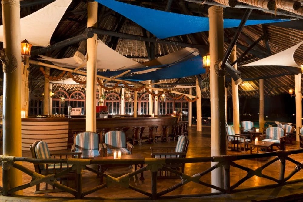 Bar all'aperto del Neptune Paradise Beach Resort con poltrone a righe, tavoli in legno e illuminazione d'ambiente sotto un tetto di paglia. Vele di tessuto bianche e blu pendono dal soffitto, creando un'atmosfera rilassata e invitante tipica dei migliori resort sulla spiaggia in Kenya.
