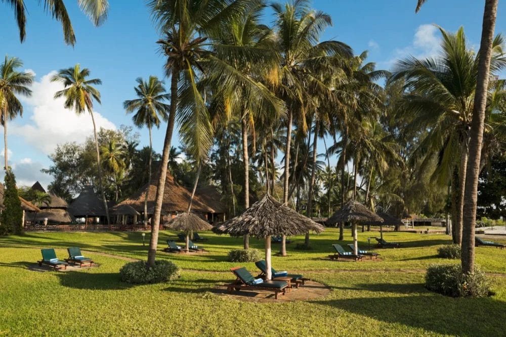 Giardino tropicale del Neptune Paradise Beach Resort in Kenya, con palme, ombrelloni di paglia, sedie a sdraio blu e un prato erboso sotto un cielo limpido. Sullo sfondo sono visibili le strutture termali e le incantevoli capanne.